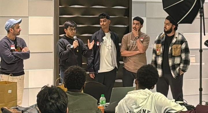 Four people standing at the front of a room, one speaking into a microphone, presenting to a seated audience.