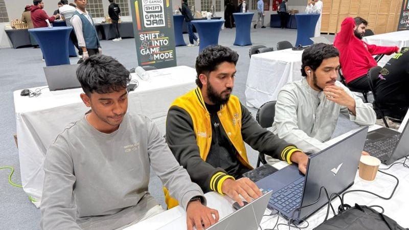 Three people working on laptops at a long table, under a SHIFT Game Jam sign reading 'shift into game mode'.