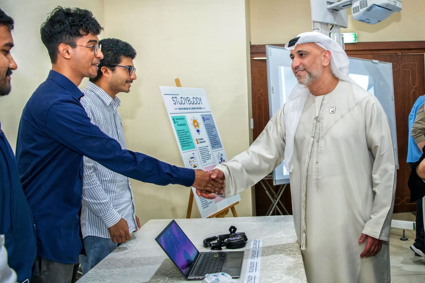 Two people shaking hands across a demo table carrying a laptop and a StudyBuddy project poster.